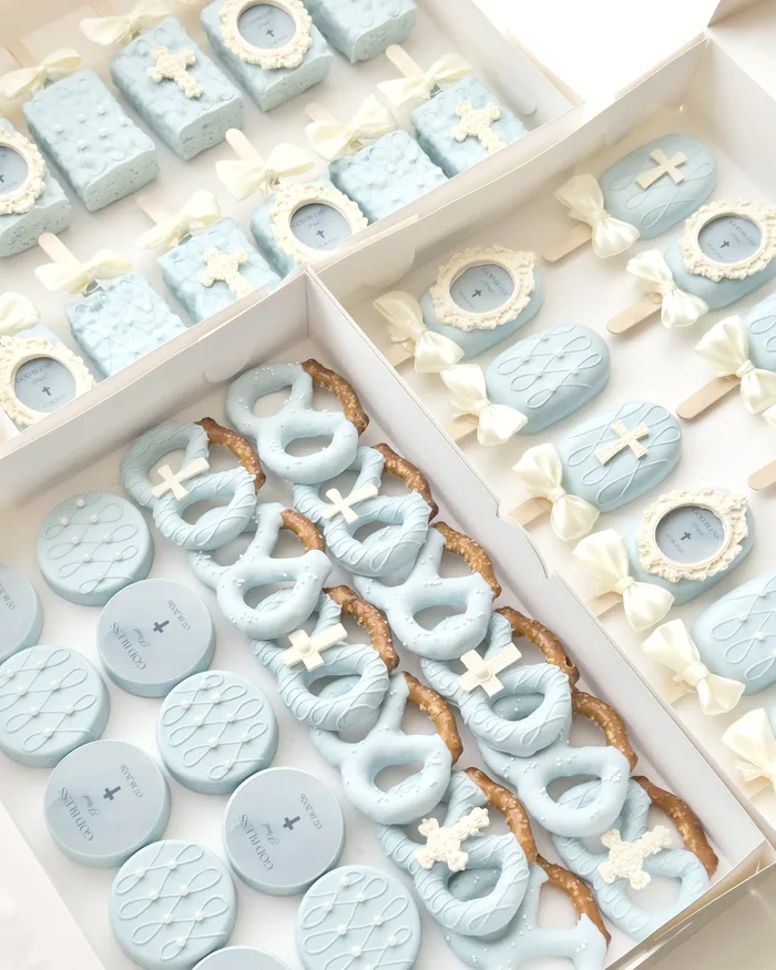 Open boxes of pale blue baptism treats: cakesicles, dessert bars, covered Oreos, and dipped pretzels with sugar crosses.