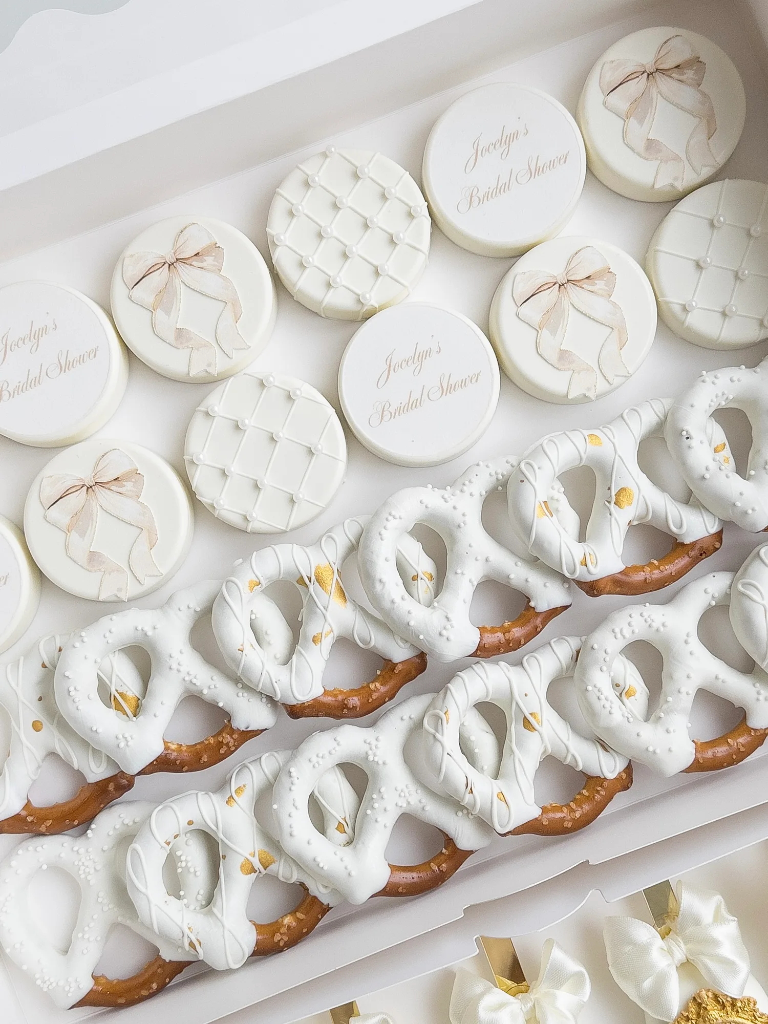 Ivory covered Oreos printed with a bridal shower name and tied with small bows, beside white dipped pretzels.