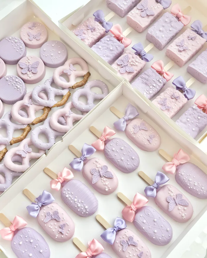 Open boxes of lavender and pink butterfly treats: cakesicles, dessert bars, covered Oreos, and dipped pretzels.