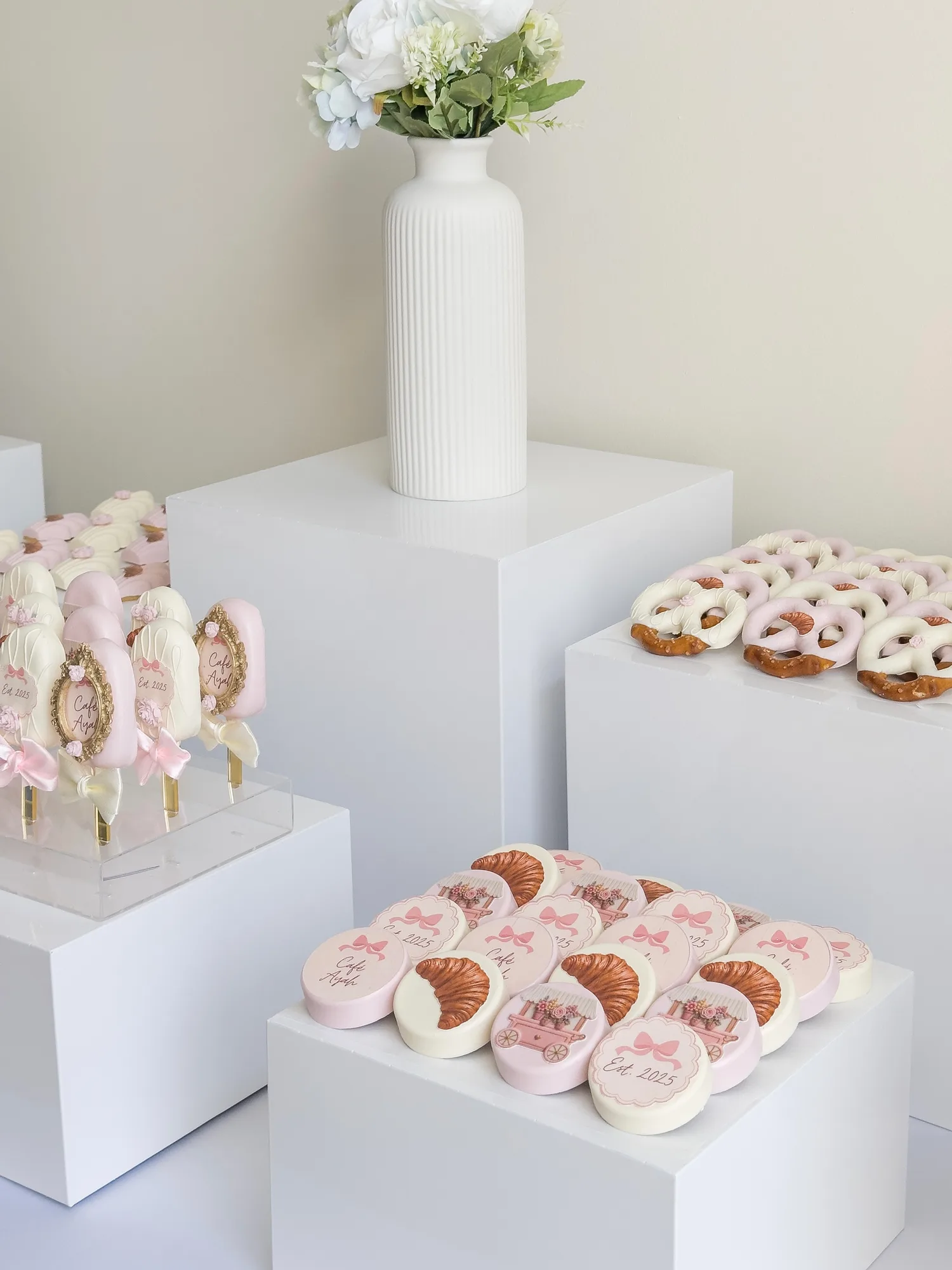 Chocolate covered Oreos printed with a cafe name, croissants, and pink bows, beside pink and white chocolate covered pretzels.