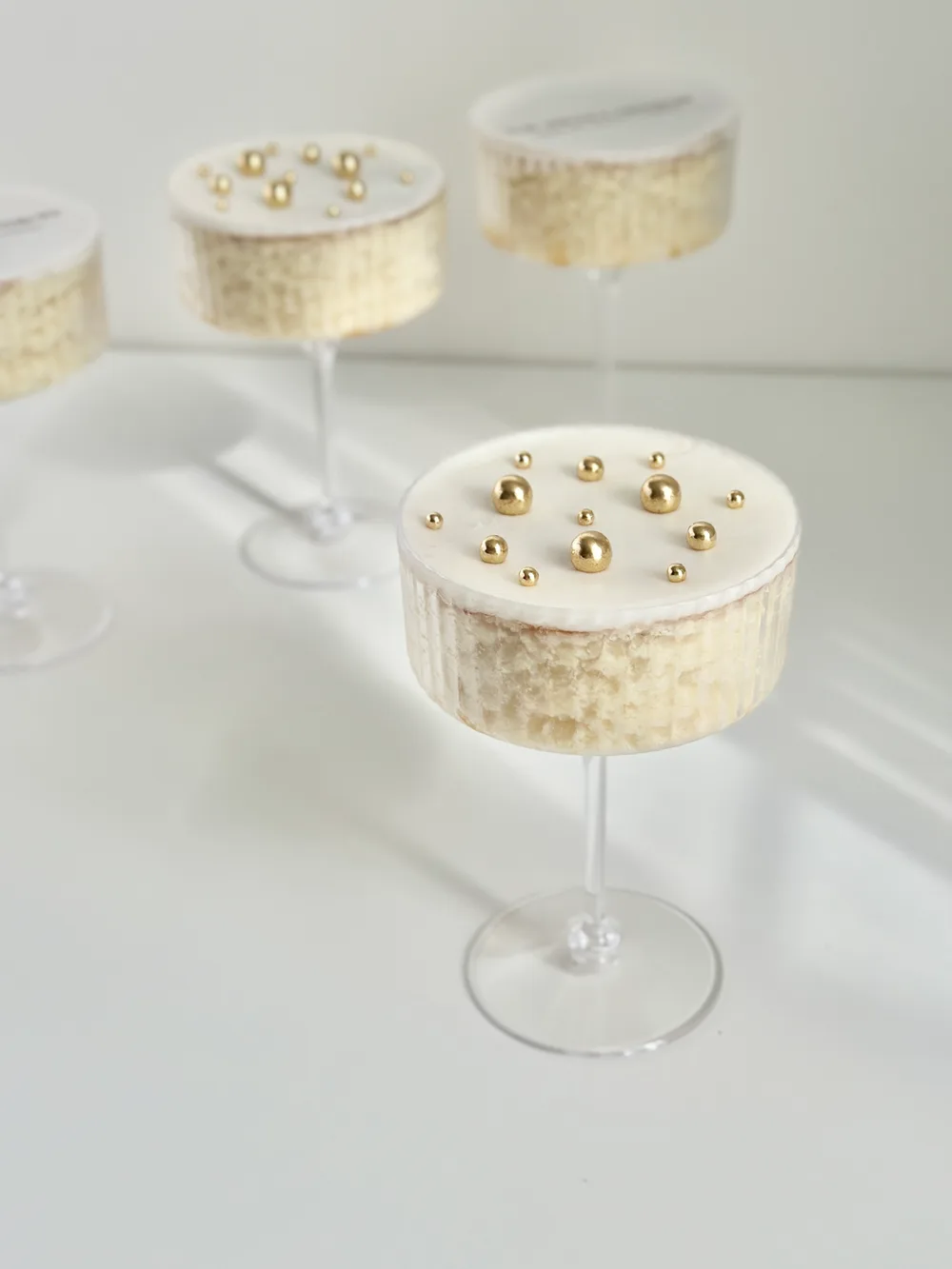A vanilla cake martini in a ribbed coupe glass, topped with white fondant and gold sugar pearls, with more cake martinis behind it.