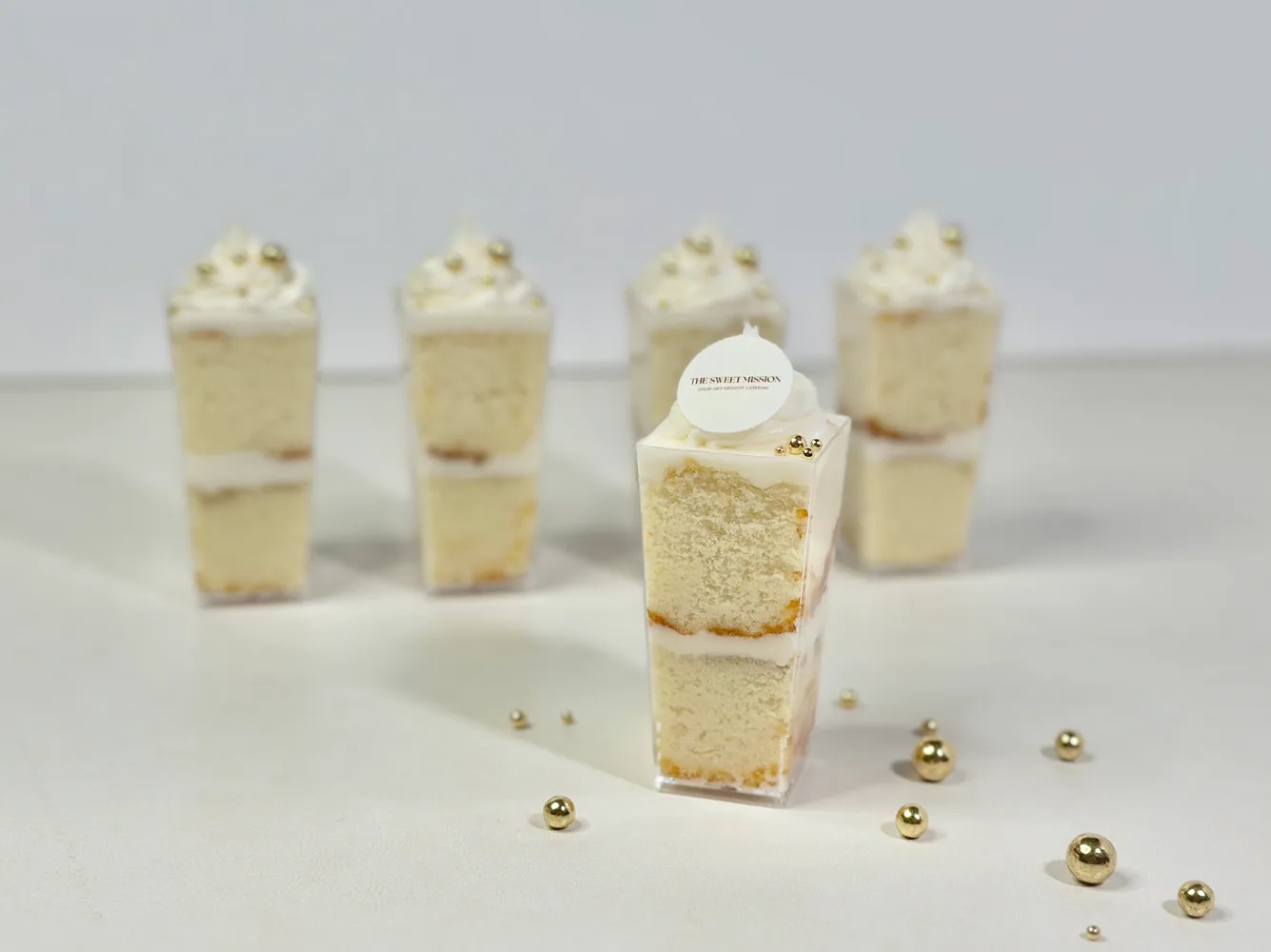 A vanilla cake shooter in a clear cup, topped with buttercream, gold sugar pearls, and a round plaque printed with The Sweet Mission logo.