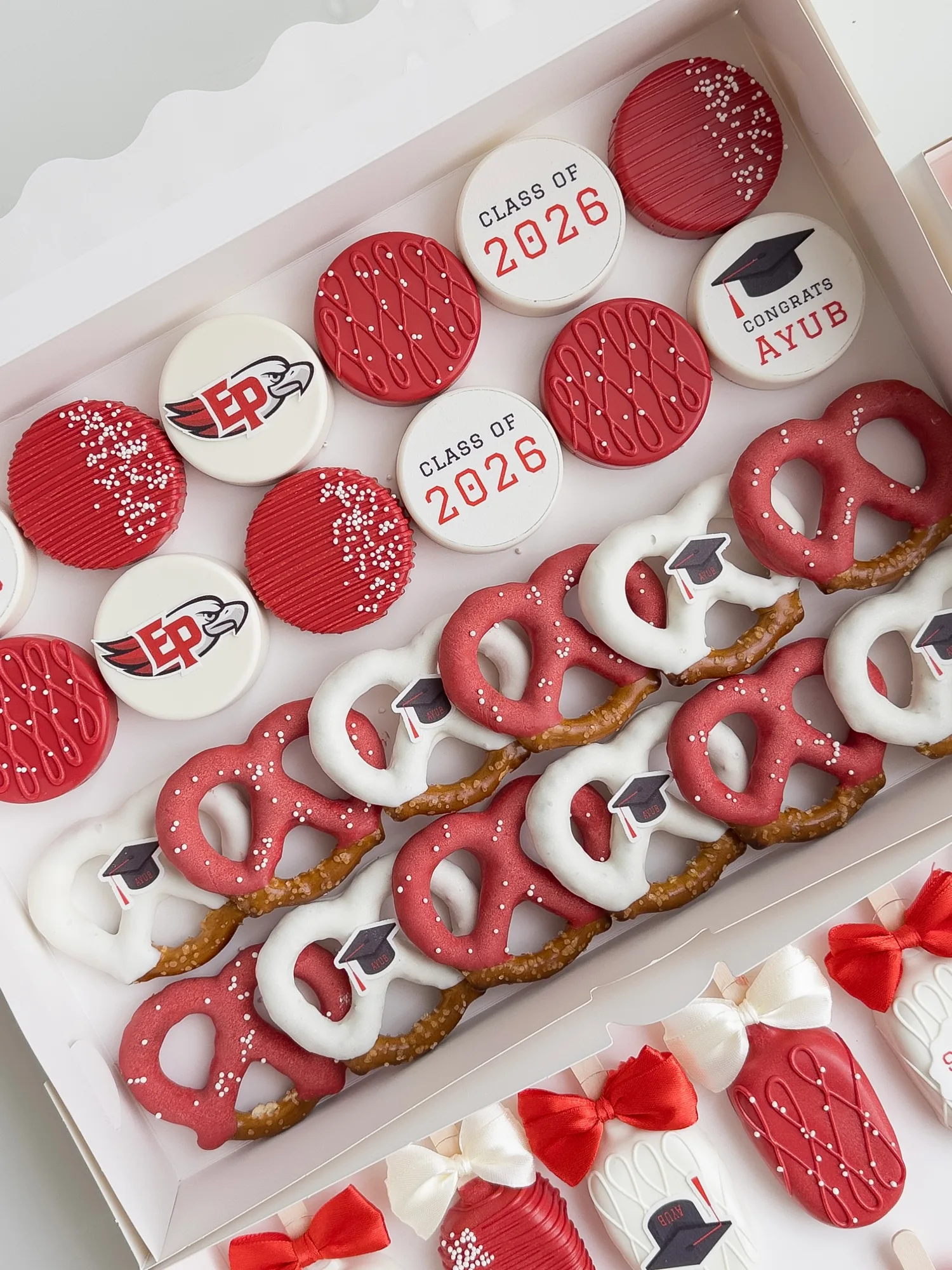 Red and white graduation treats: covered Oreos printed with a school logo and Class of 2026, beside dipped pretzels with sugar graduation caps.