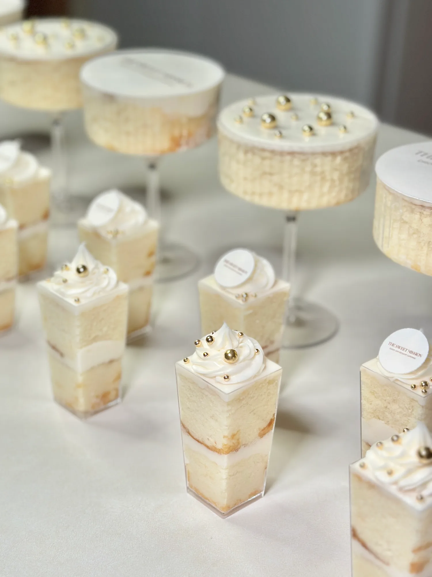 Vanilla cake shooters and cake martinis with white buttercream, gold sugar pearls, and round edible plaques printed with The Sweet Mission logo.