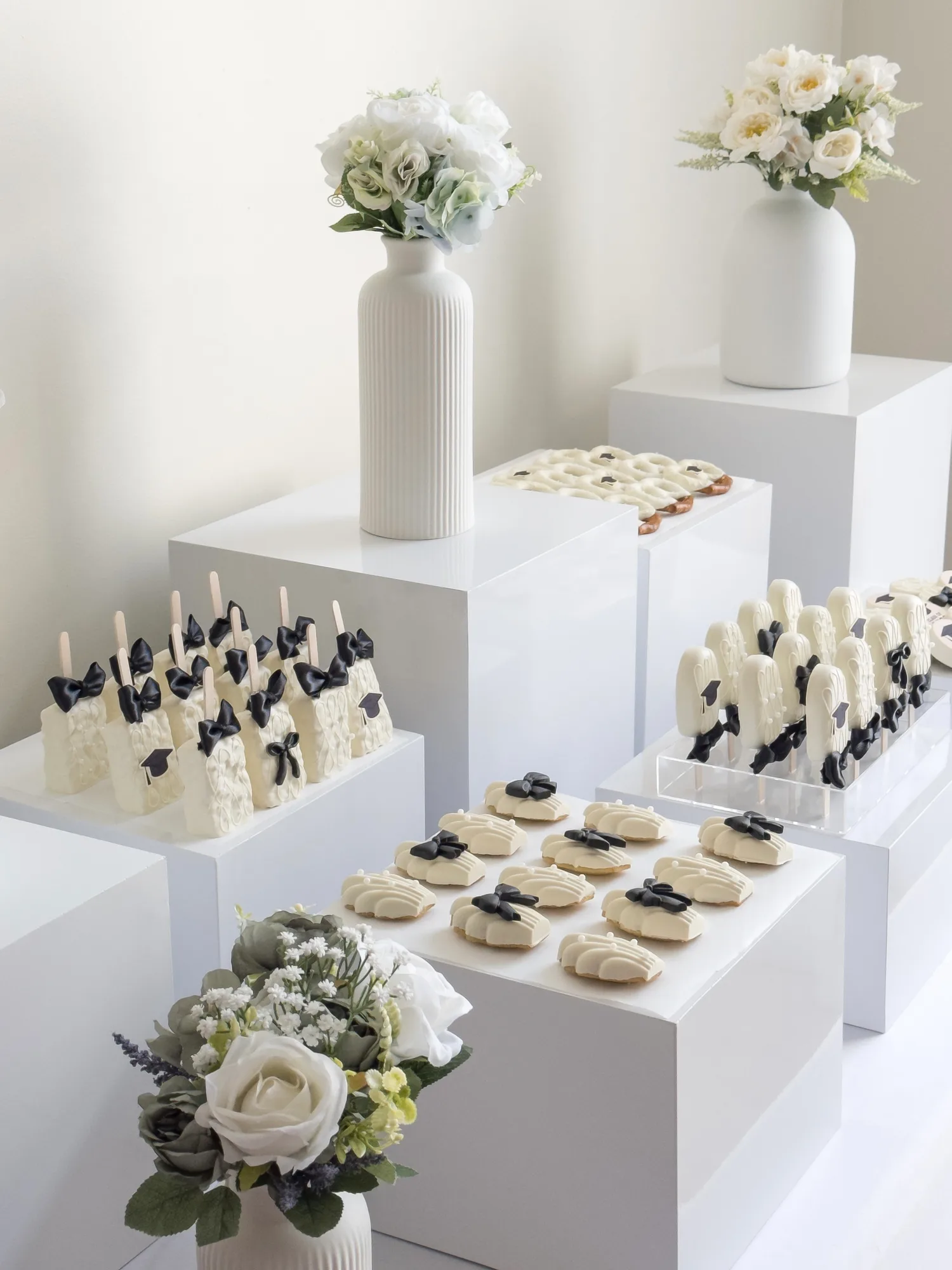 A white graduation dessert display with ivory chocolate treats finished in black satin bows and sugar graduation caps, beside vases of white flowers.
