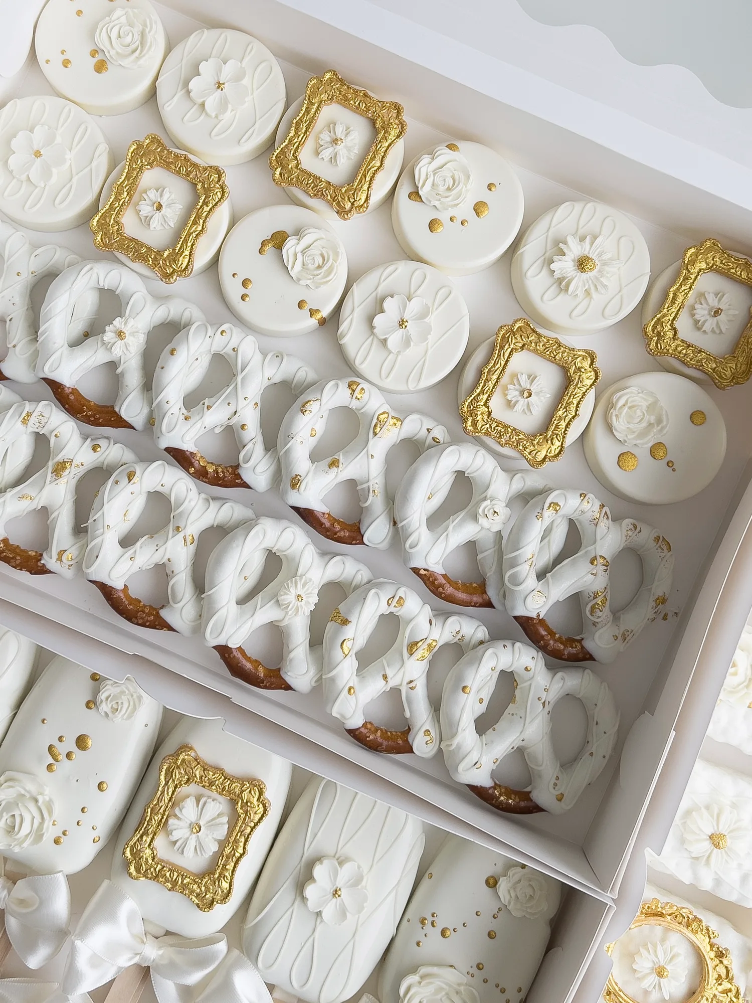 An open white box of all ivory covered Oreos with gold framed plaques and sugar flowers, beside white dipped pretzels flecked with gold.