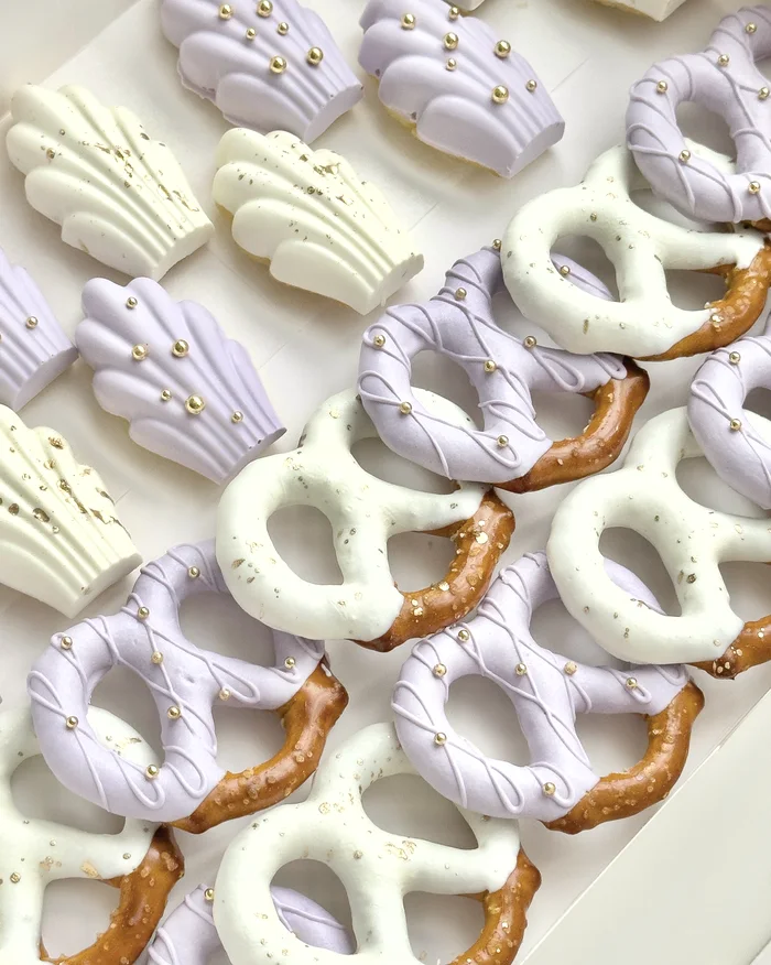A box of lavender and ivory madeleines and chocolate dipped pretzels, flecked with gold sugar pearls.