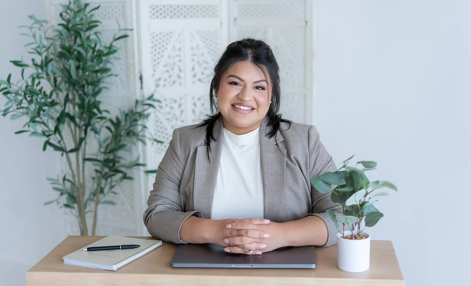 Maria, the owner of The Sweet Mission, seated at a wood desk in a taupe blazer with her hands folded on a closed laptop, a notebook and pen to one side and a small eucalyptus plant to the other.
