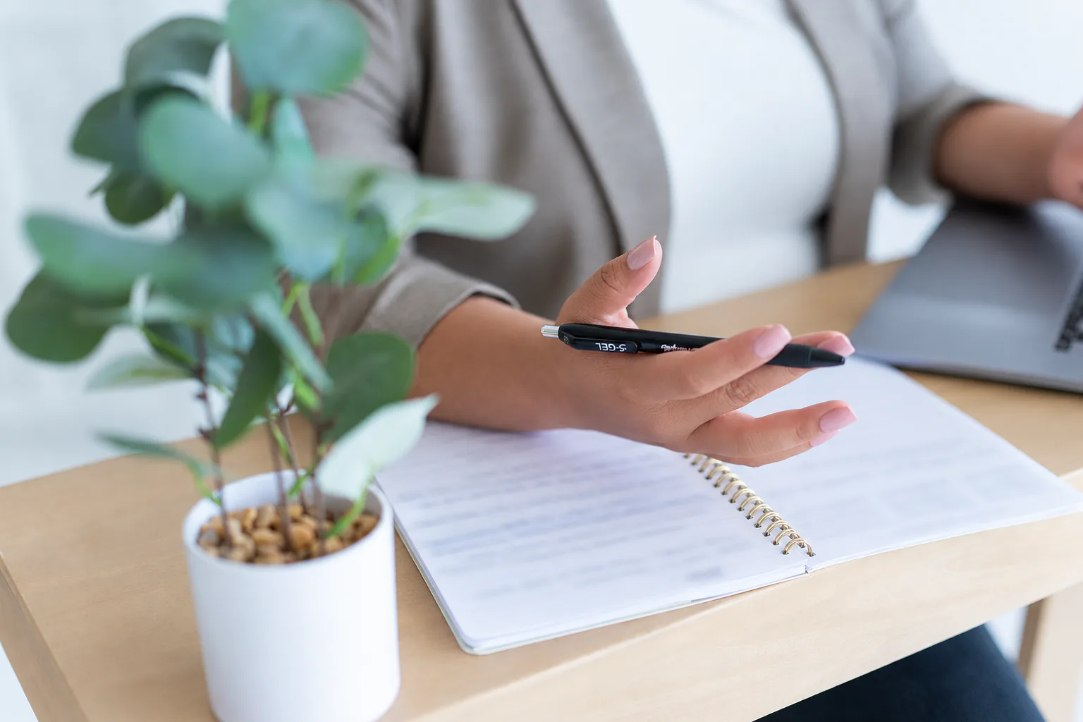 Maria's hand holding a pen over an open planner, a small eucalyptus plant beside it on a wood desk.