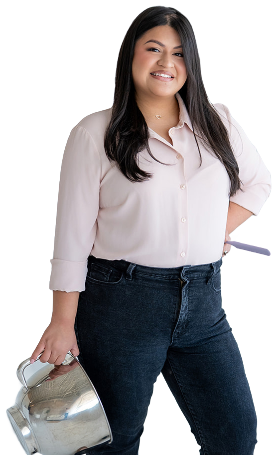 Maria, the owner of The Sweet Mission, stands in a blush blouse and dark jeans, smiling, with a steel mixing bowl in one hand and a spatula in the other.