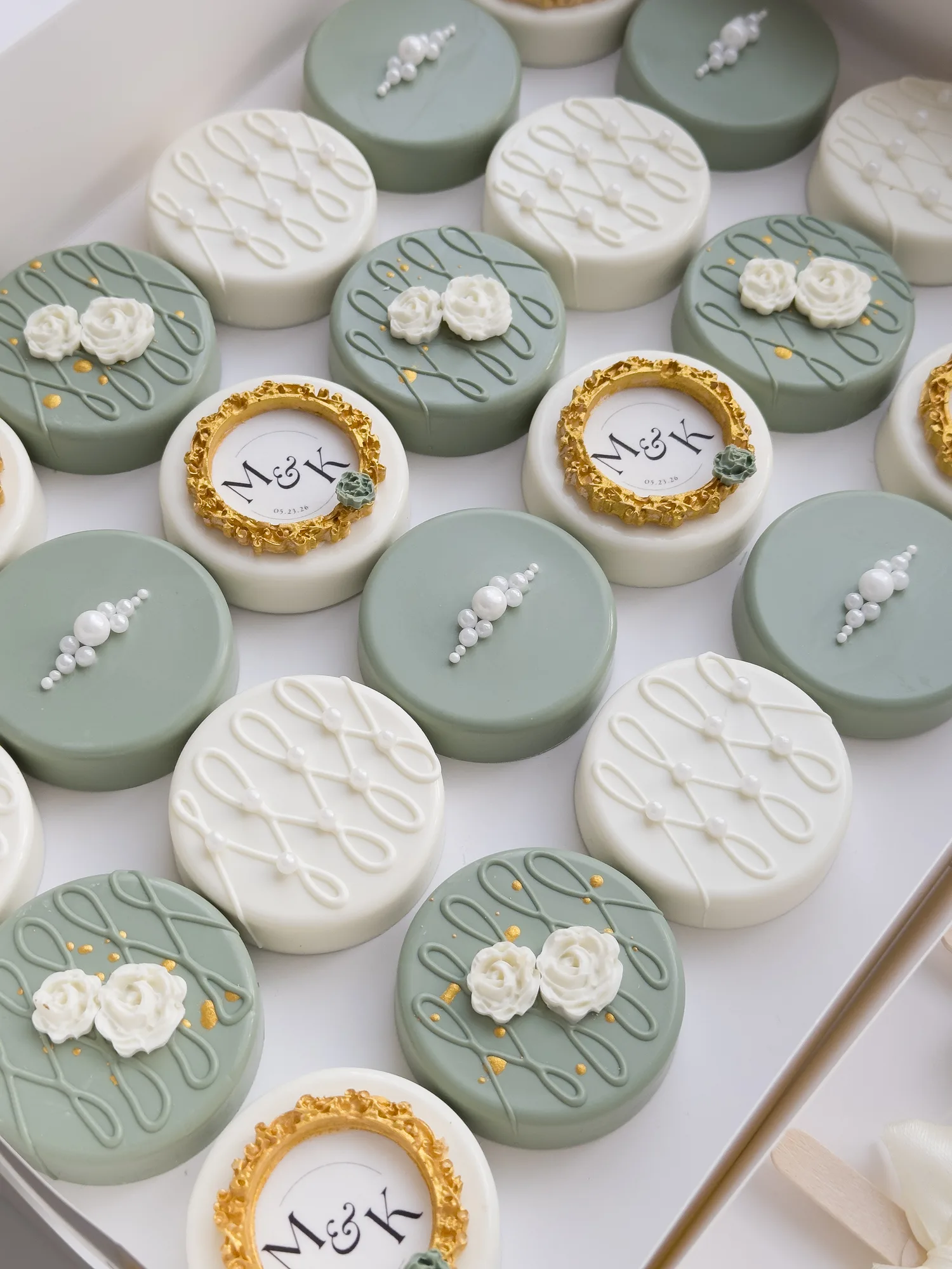 A close view of sage green and ivory covered Oreos with gold framed monogram plaques and piped roses.