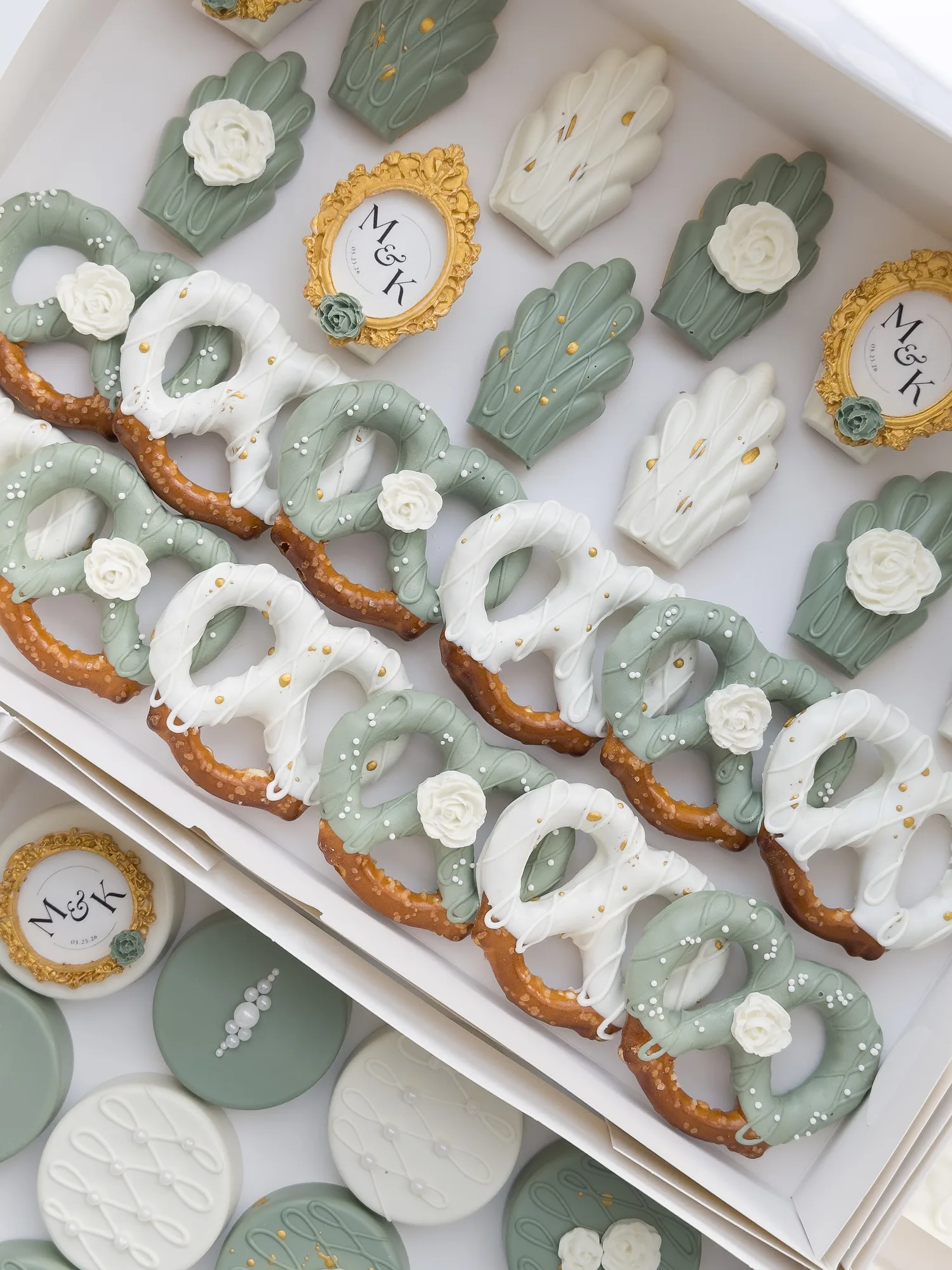 A white box of sage green and ivory treats: covered Oreos with gold monogram frames, dipped madeleines, and dipped pretzels.