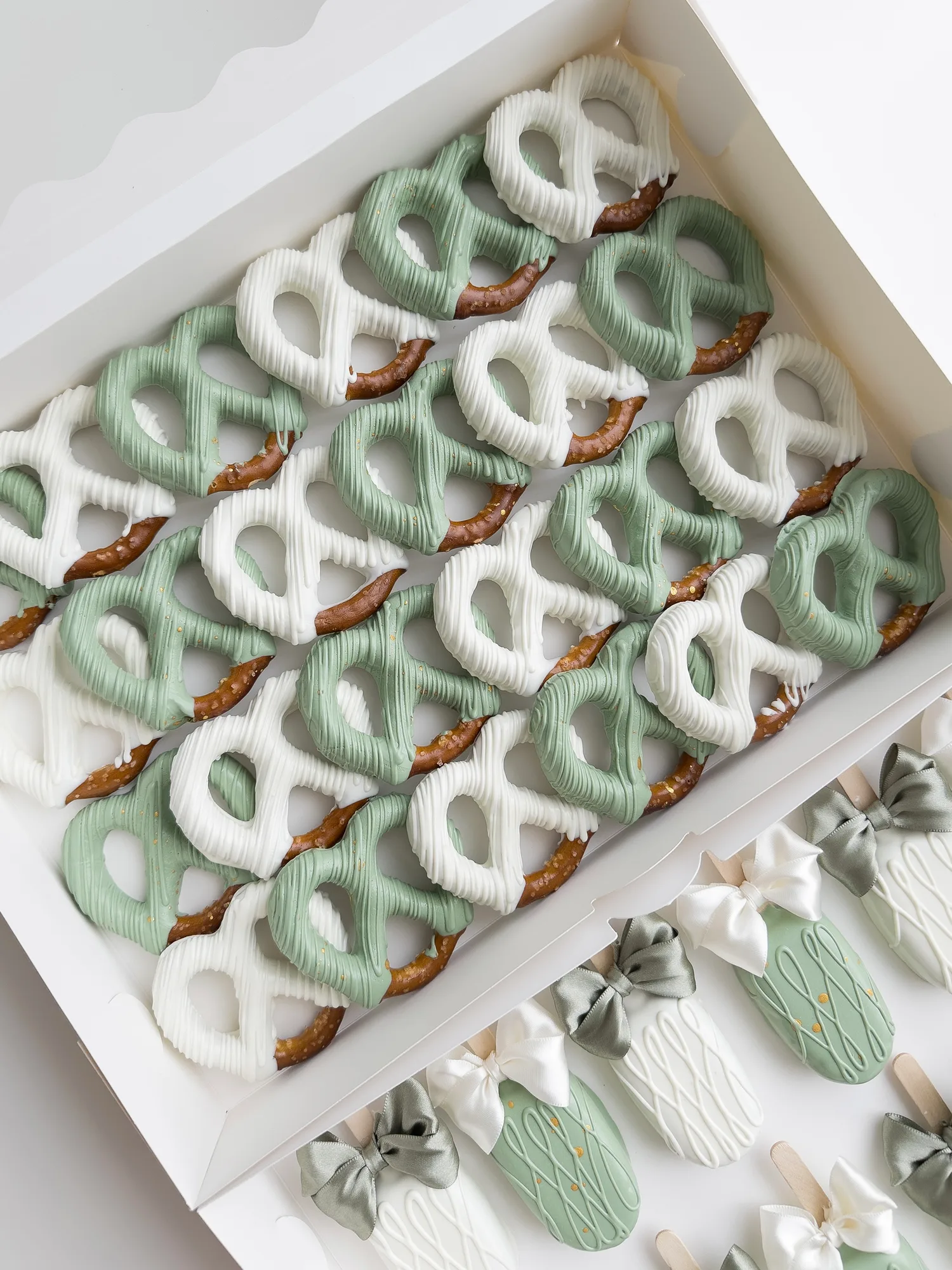 Rows of sage green and white chocolate dipped pretzels with small bows, packed in white boxes for a large order.
