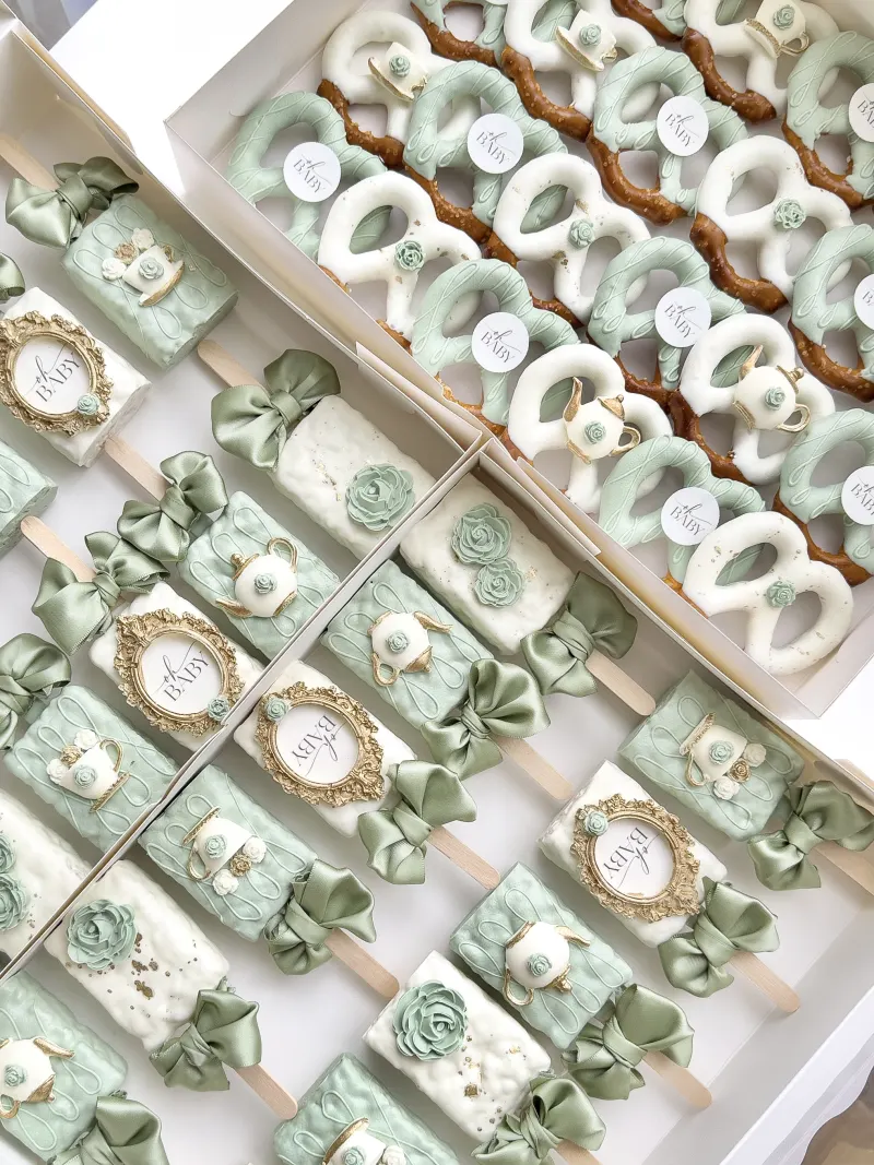 Sage green and ivory cakesicles with gold framed oh baby plaques, sugar teapots, and sage satin bows, beside a box of matching pretzels.