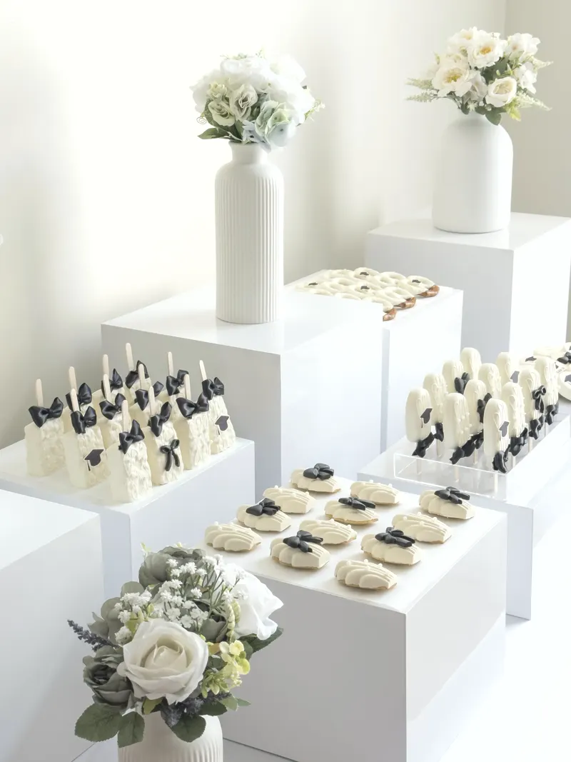 A white pedestal display of graduation desserts in ivory chocolate, finished with black satin bows and sugar graduation caps.