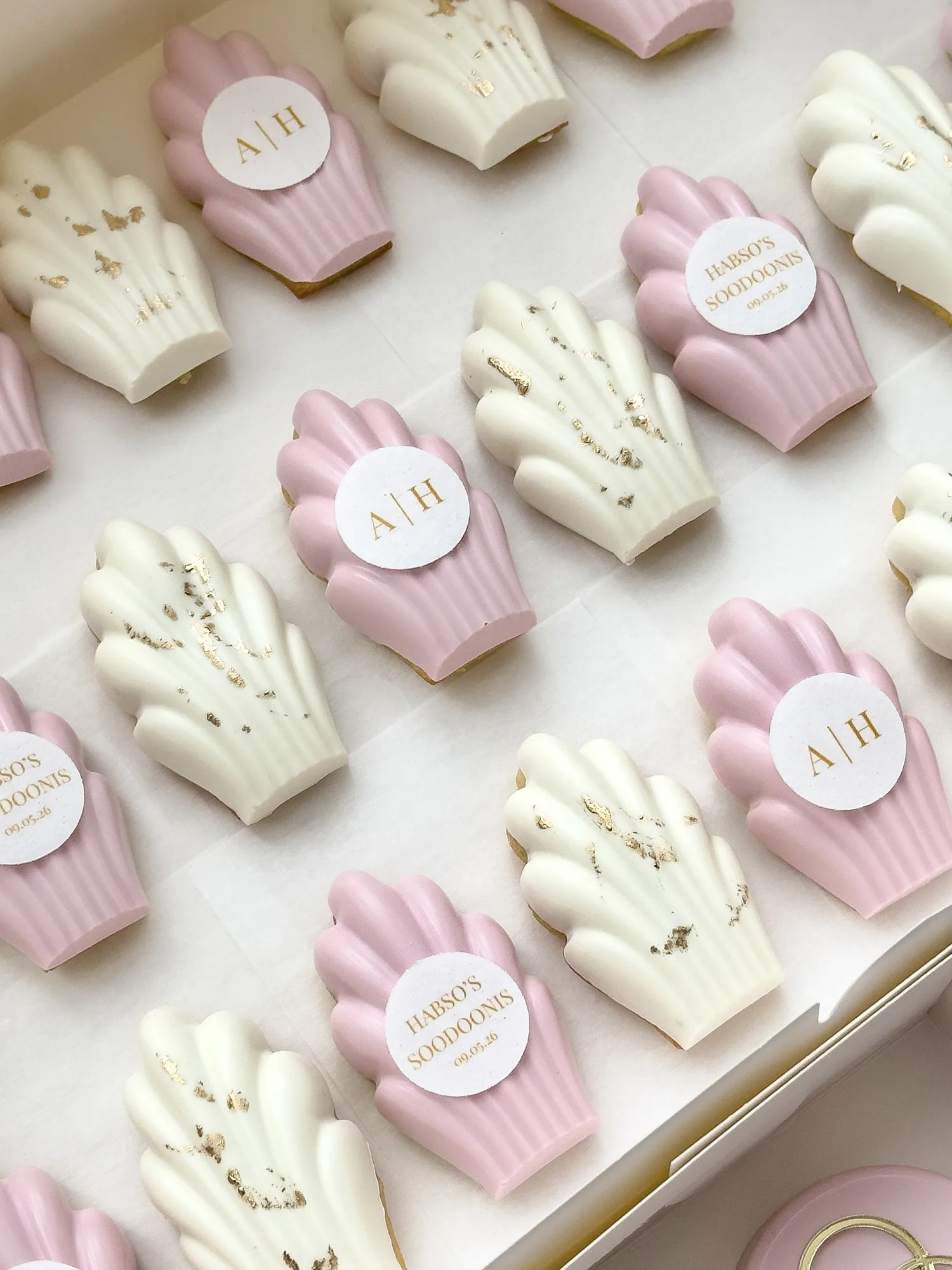 Pink and ivory dipped madeleines in a white box, some flecked with gold leaf, others carrying round monogram and name plaques.