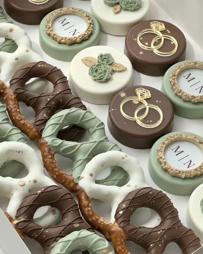 Sage and chocolate covered Oreos with gold wedding rings, piped roses, and monogram plaques, beside dipped pretzels.